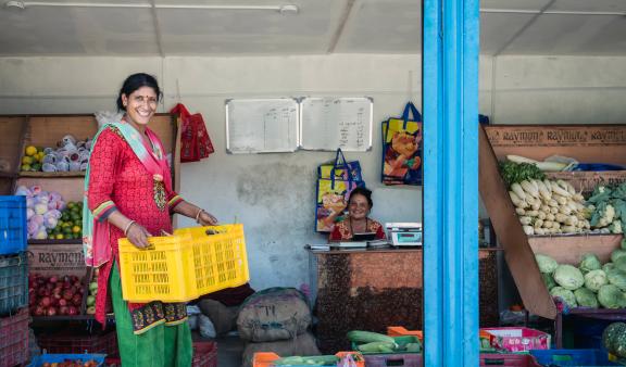 Photo of a woman setting up a vegetable stand, while another woman is sitting at the stand. Both women are smiling.