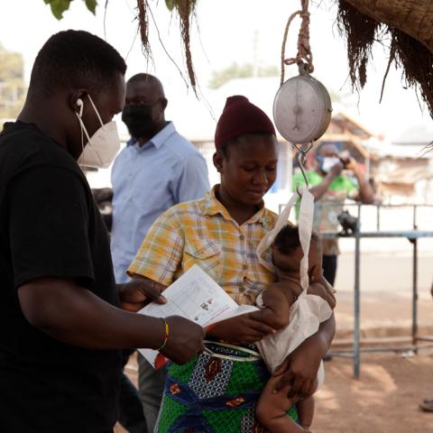 Health worker documenting small infant's weight outside with a hanging scale, while the mother holding the child.