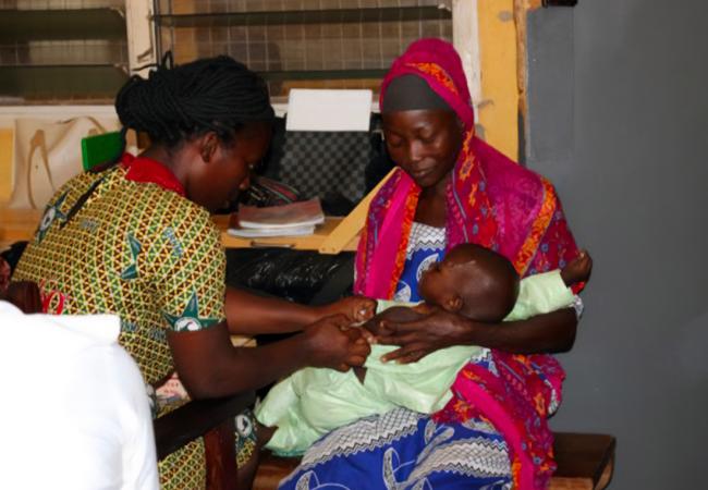 Woman sitting in her house, holding her young infant, as a health worker does a physical check of the child.